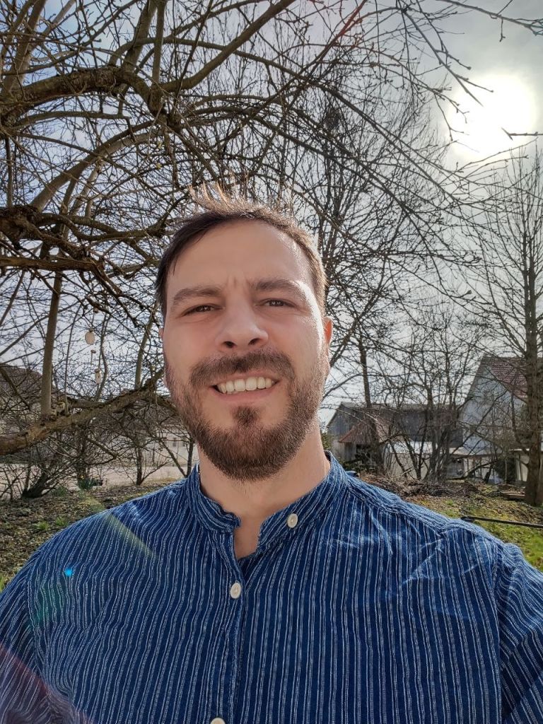 Carl Zipser, lächelnd, im blau gestreiften Hemd unter kahlen Obstbäumen.